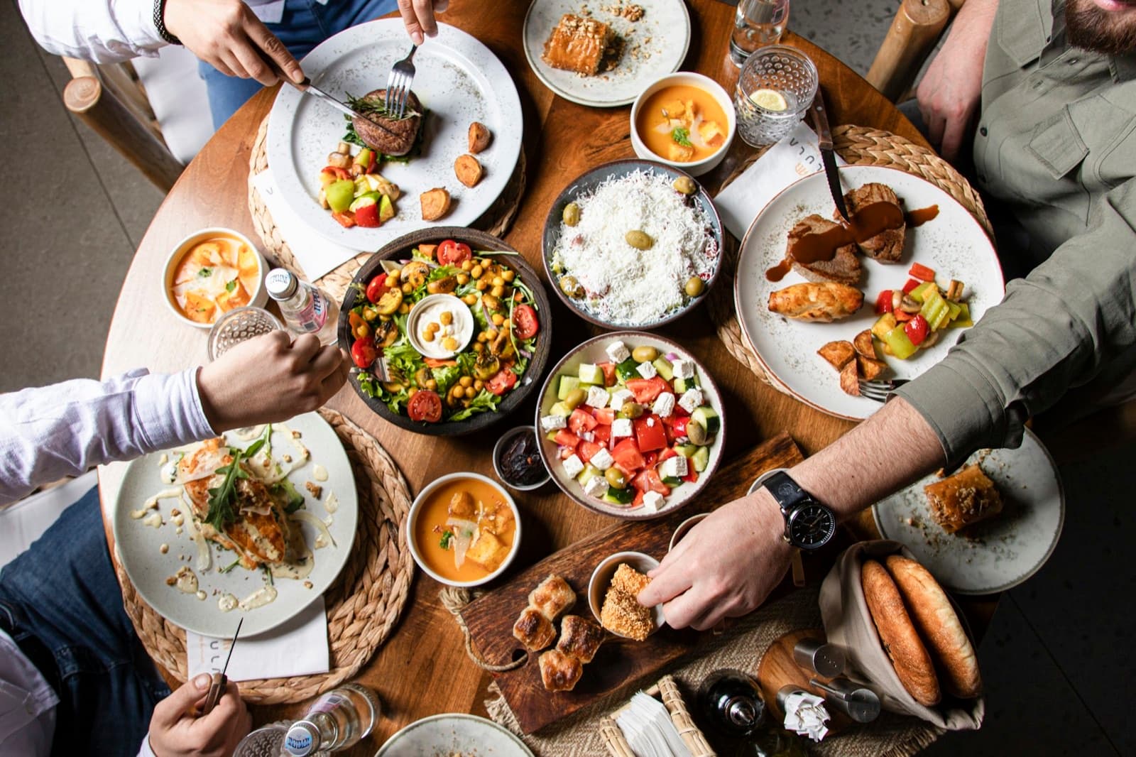 Gedeckter Restauranttisch von oben mit mehreren Tellern, Gläsern und Händen, die sich Speisen teilen