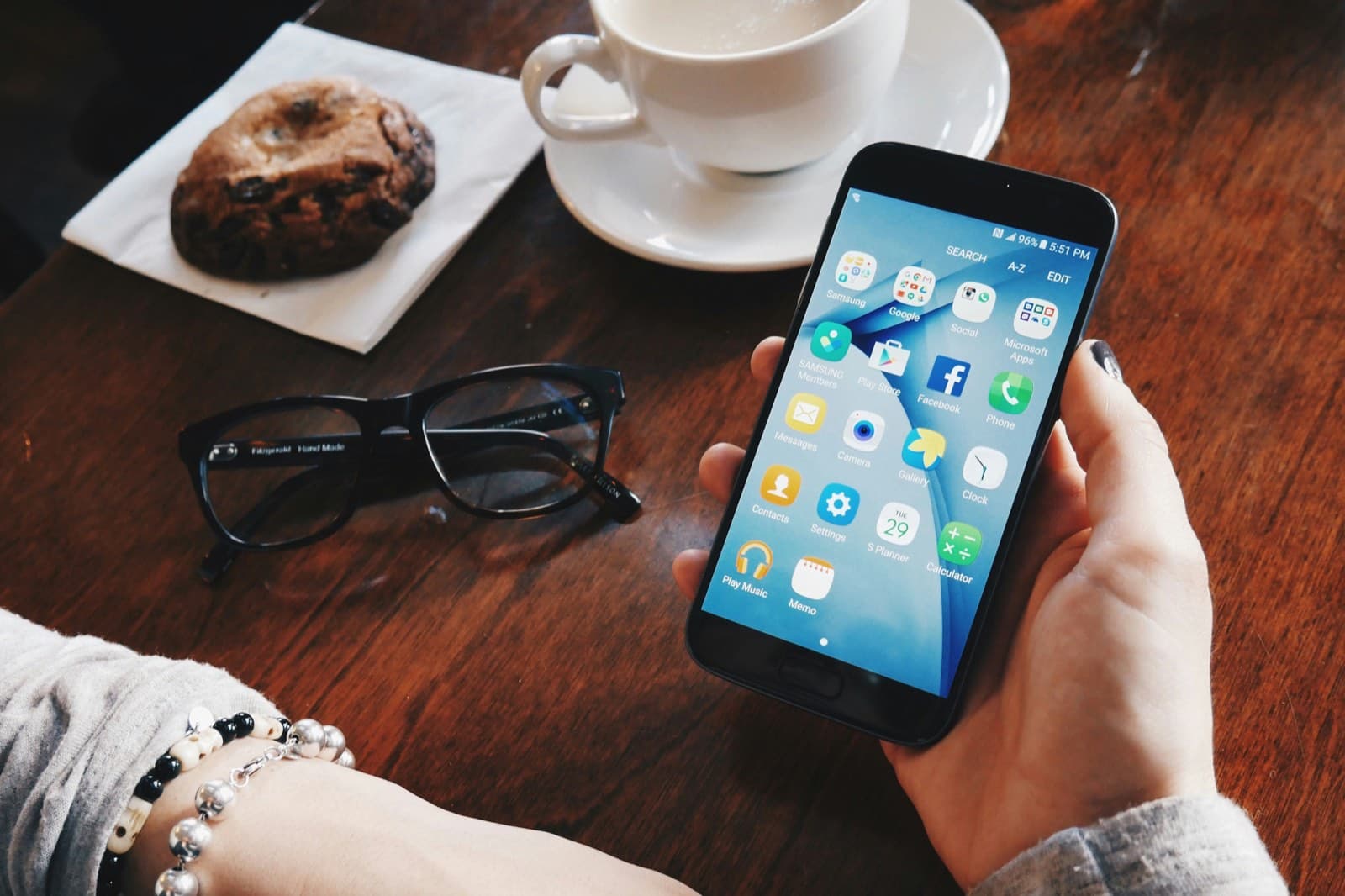 Hand hält ein Smartphone mit geöffneter App-Übersicht über einem Cafétisch mit Kaffeetasse und Brille