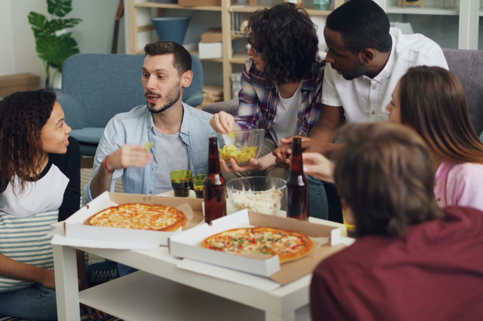 Sechs Freunde sitzen auf dem Sofa und dem Boden um einen Couchtisch mit zwei gelieferten Pizzen in offenen Kartons, Bier und Chips