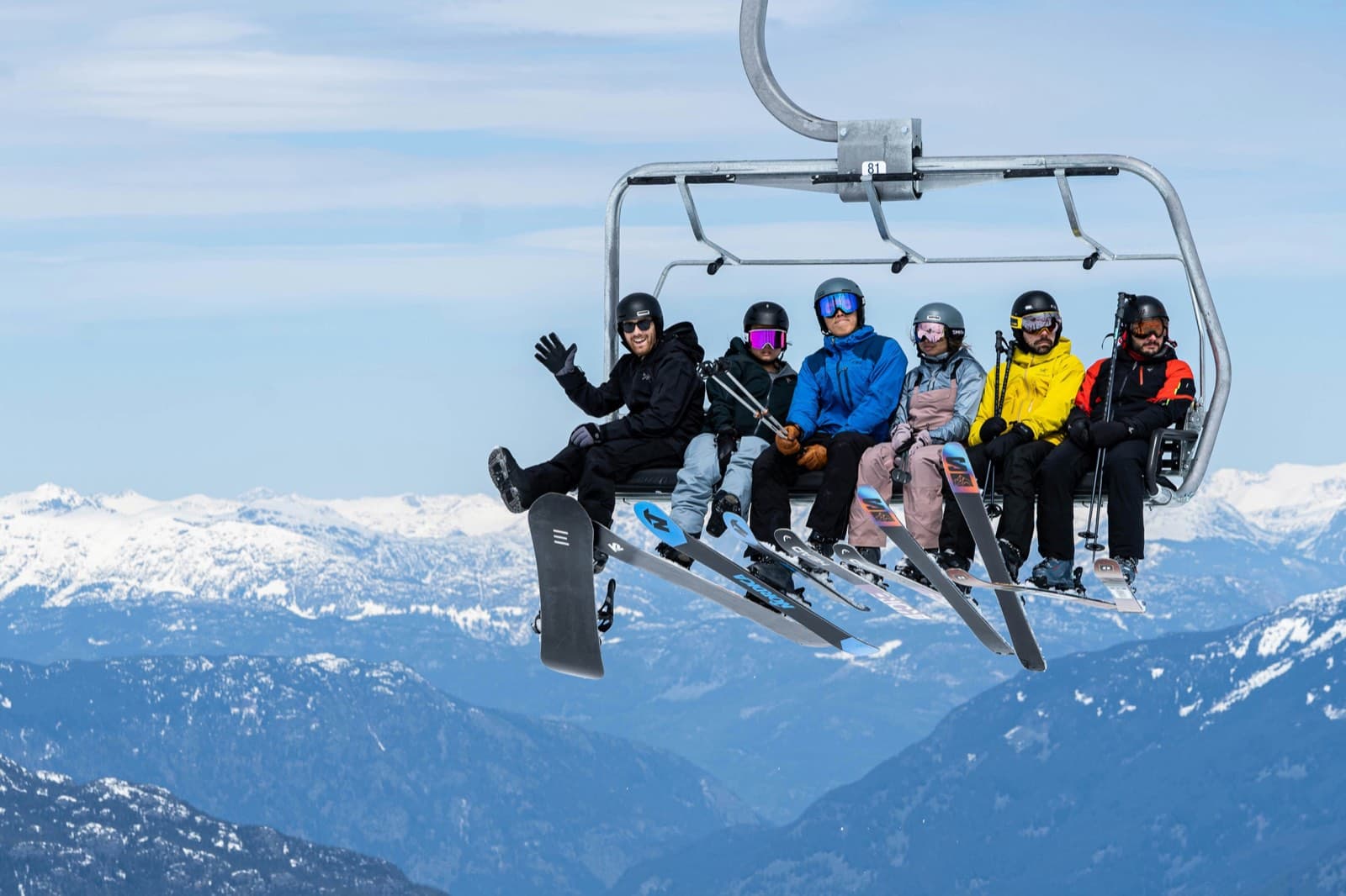 Sechs Freunde mit Skiern und Snowboard sitzen gemeinsam im Sessellift vor verschneiten Bergen